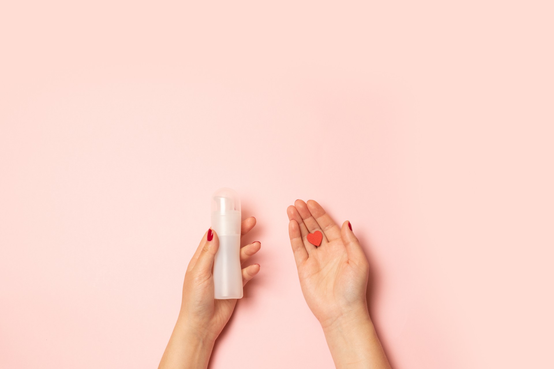 Womens hands hold intimate grease and a red heart shape on a pink background